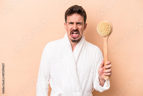 Young caucasian man holding back scratcher isolated on beige background screaming very angry and aggressive.