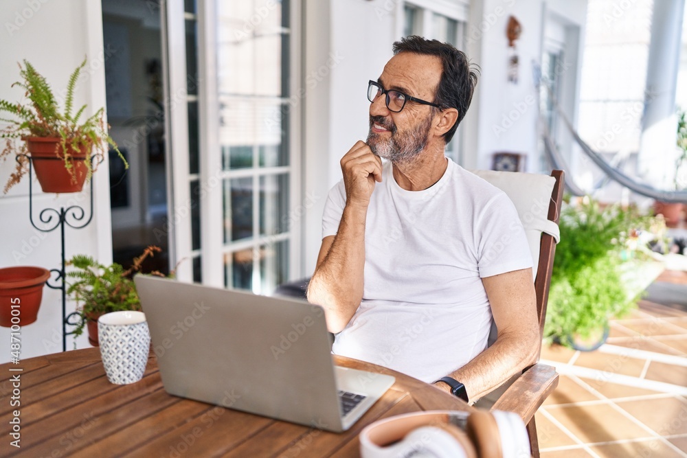 Middle age man using computer laptop at home with hand on chin thinking about question, pensive expression. smiling with thoughtful face. doubt concept.