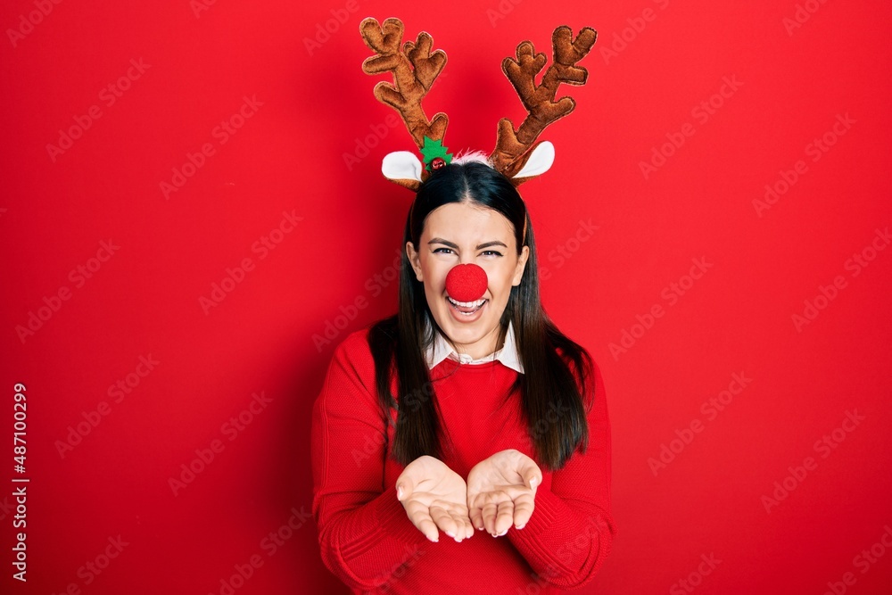 Young hispanic woman wearing deer christmas hat and red nose smiling with hands palms together receiving or giving gesture. hold and protection Young hispanic woman wearing deer christmas hat and red nose smiling with hands palms together receiving or giving gesture. hold and protection