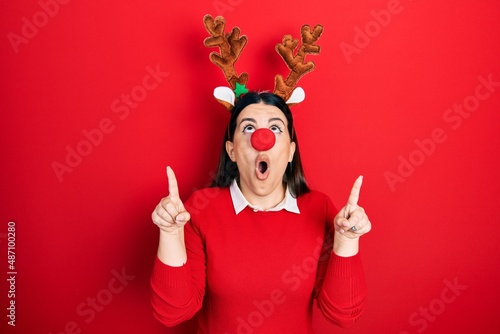 Young hispanic woman wearing deer christmas hat and red nose amazed and surprised looking up and pointing with fingers and raised arms.