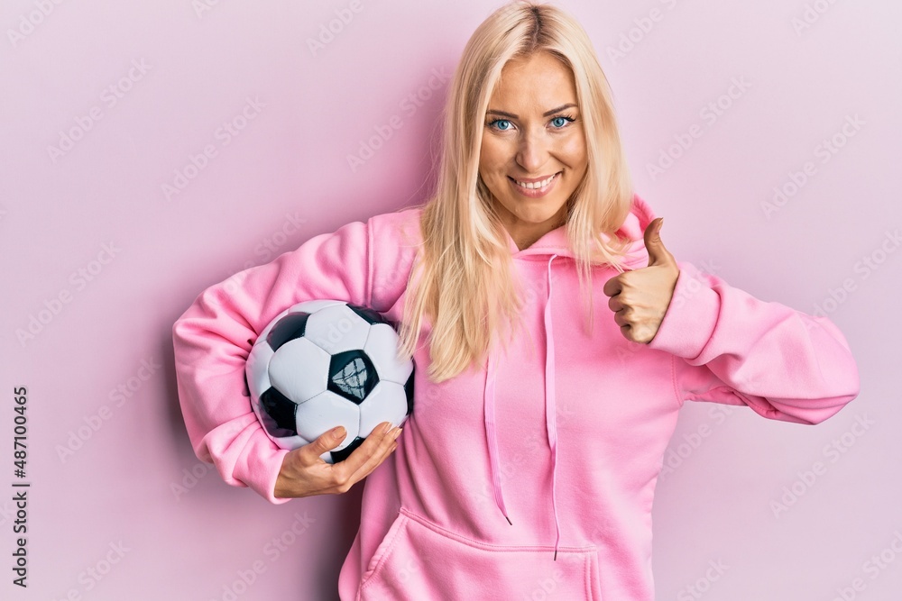 Young blonde woman holding soccer ball smiling happy and positive, thumb up doing excellent and approval sign Young blonde woman holding soccer ball smiling happy and positive, thumb up doing excellent and approval sign