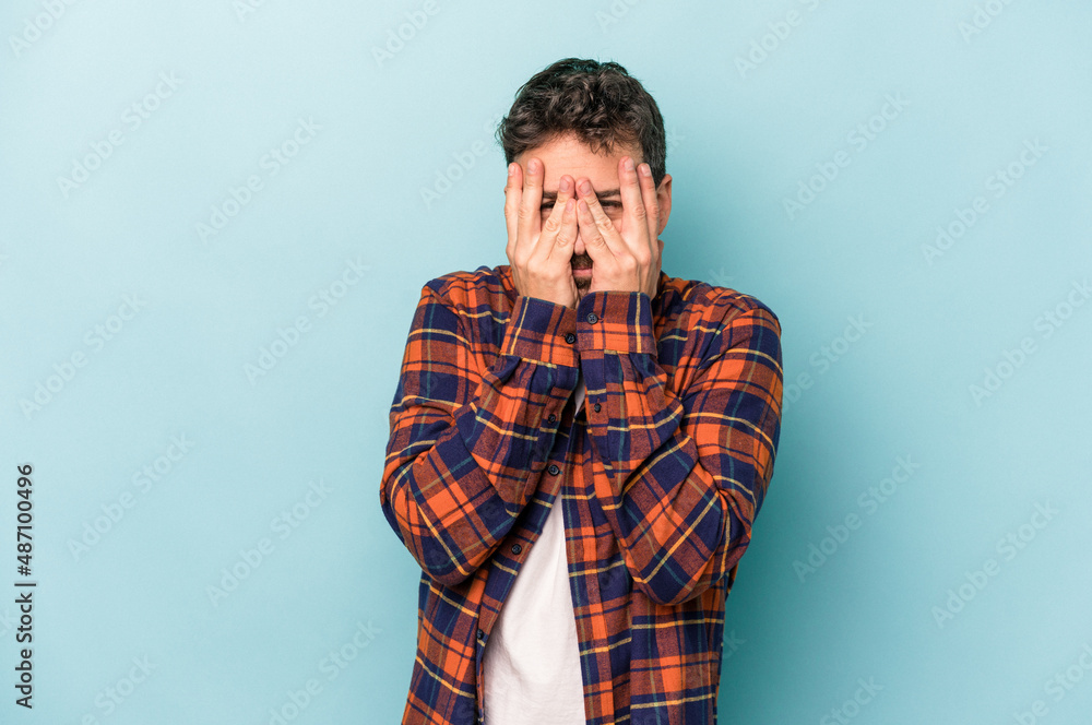 Fototapeta premium Young caucasian man isolated on blue background blink at the camera through fingers, embarrassed covering face.