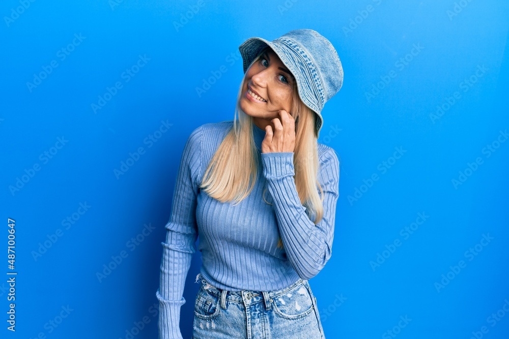 Young blonde woman wearing casual denim hat smiling with hand over ear listening an hearing to rumor or gossip. deafness concept.