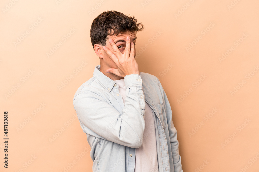 Young caucasian man isolated on beige background blink at the camera through fingers, embarrassed covering face. Young caucasian man isolated on beige background blink at the camera through fingers, embarrassed covering face.