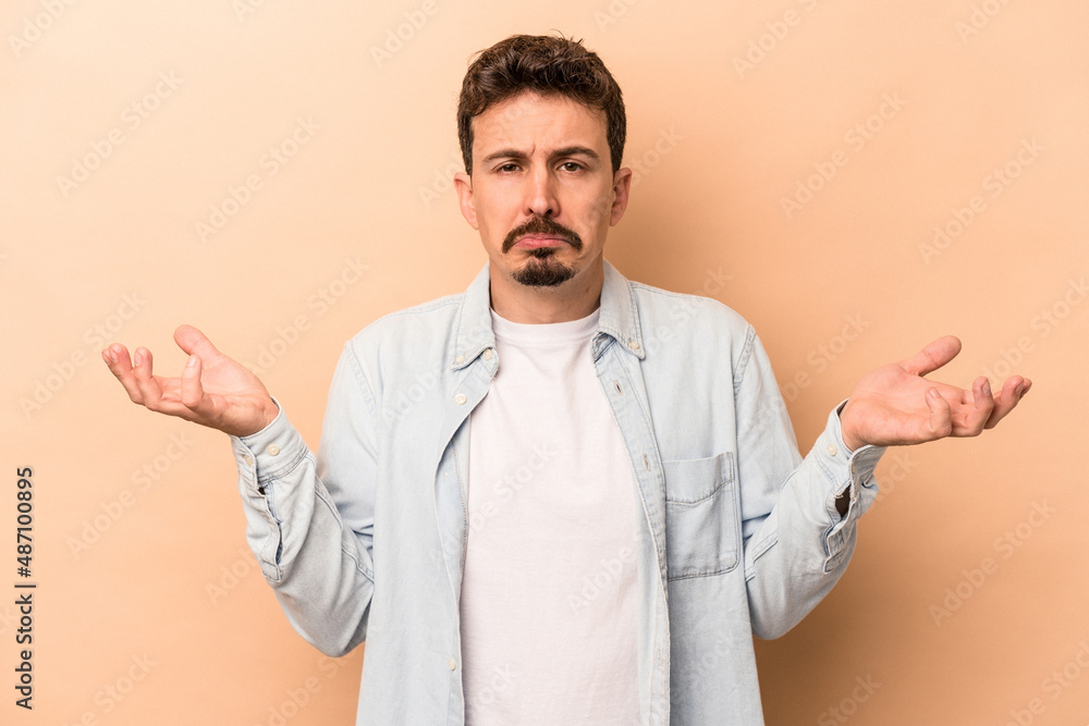 Young caucasian man isolated on beige background doubting and shrugging shoulders in questioning gesture. Young caucasian man isolated on beige background doubting and shrugging shoulders in questioning gesture.