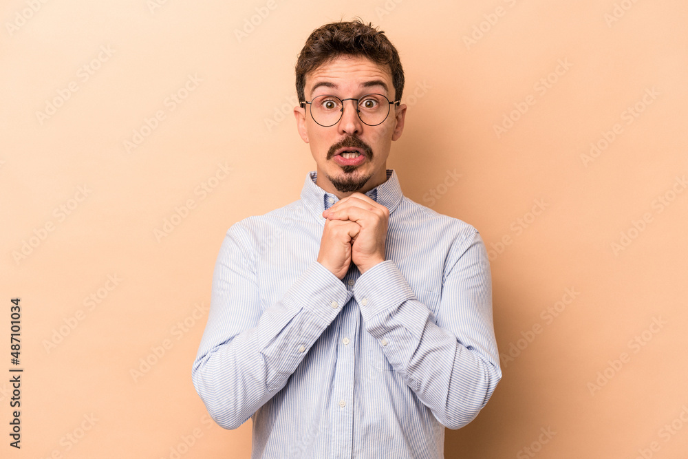Young caucasian man isolated on beige background praying for luck, amazed and opening mouth looking to front.