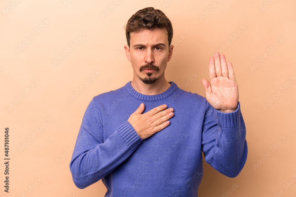 Young caucasian man isolated on beige background taking an oath, putting hand on chest. Young caucasian man isolated on beige background taking an oath, putting hand on chest.