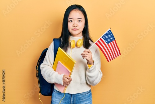Young chinese girl exchange student holding america flag relaxed with serious expression on face. simple and natural looking at the camera.