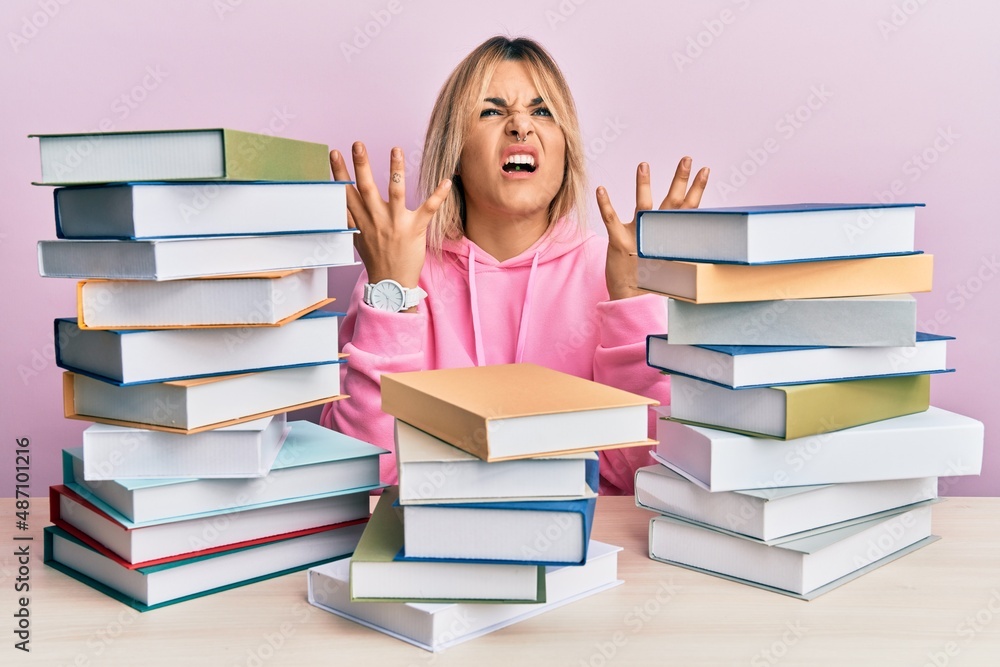 Young caucasian woman sitting on the table with books crazy and mad shouting and yelling with aggressive expression and arms raised. frustration concept. Young caucasian woman sitting on the table with books crazy and mad shouting and yelling with aggressive expression and arms raised. frustration concept.