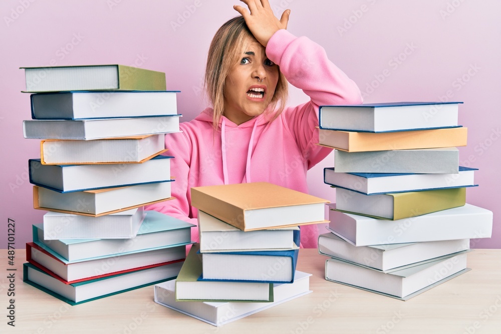 Young caucasian woman sitting on the table with books surprised with hand on head for mistake, remember error. forgot, bad memory concept. Young caucasian woman sitting on the table with books surprised with hand on head for mistake, remember error. forgot, bad memory concept.