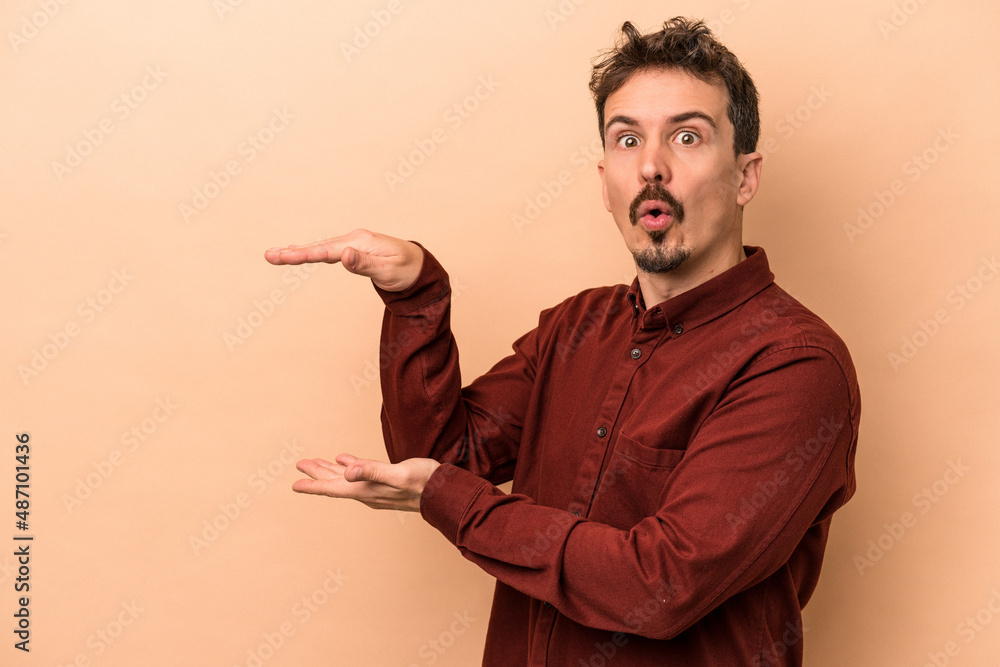 Young caucasian man isolated on beige background shocked and amazed holding a copy space between hands. Young caucasian man isolated on beige background shocked and amazed holding a copy space between hands.