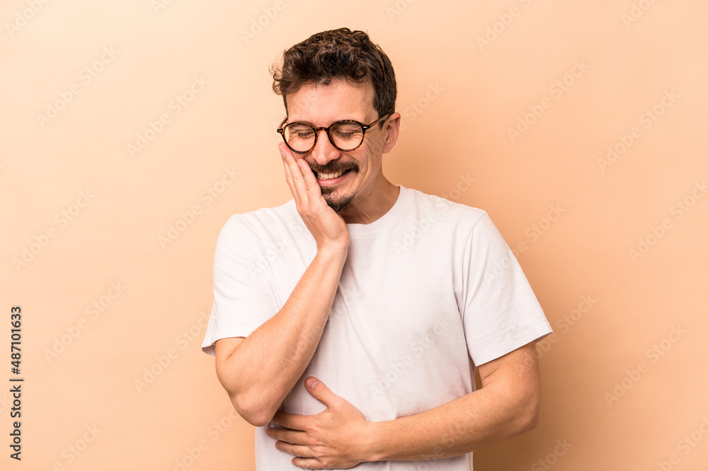 Young caucasian man isolated on beige background laughs happily and has fun keeping hands on stomach. Young caucasian man isolated on beige background laughs happily and has fun keeping hands on stomach.
