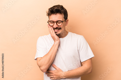 Young caucasian man isolated on beige background laughs happily and has fun keeping hands on stomach.