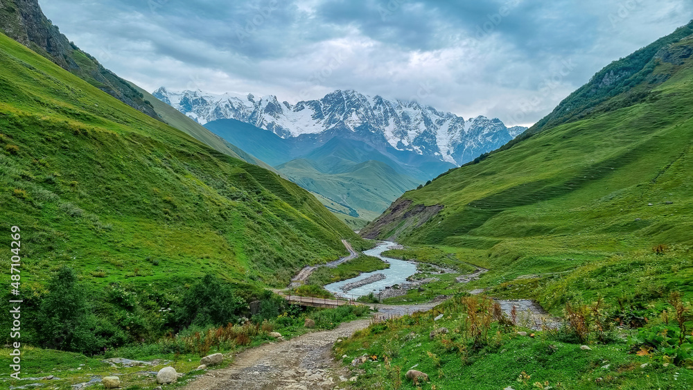 Patara Enguri River flowing down the a valley in the Greater Caucasus Mountain Range in Georgia, Svaneti Region, Ushguli. The Shkhara Mountain is covered by clouds. Snow-capped mountains.Bridge