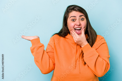 Young caucasian overweight woman isolated on blue background holds copy space on a palm, keep hand over cheek. Amazed and delighted.