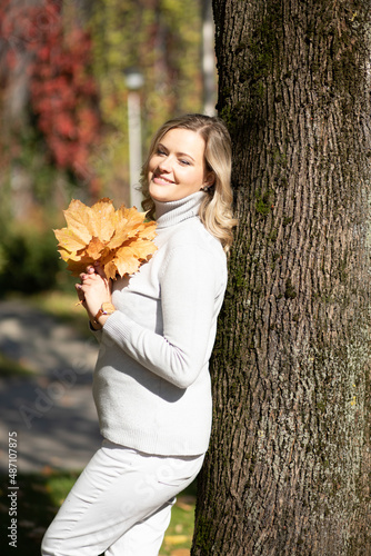 Smiling middle-aged woman with long wavy fair hair in roll-neck sweater holding bouquet of fallen leaves leaning on tree