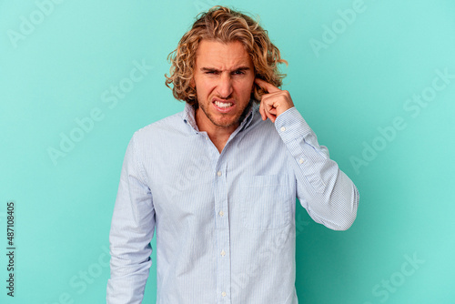 Fotografie Young caucasian man isolated on blue background covering ears with fingers, stressed and desperate by a loudly ambient