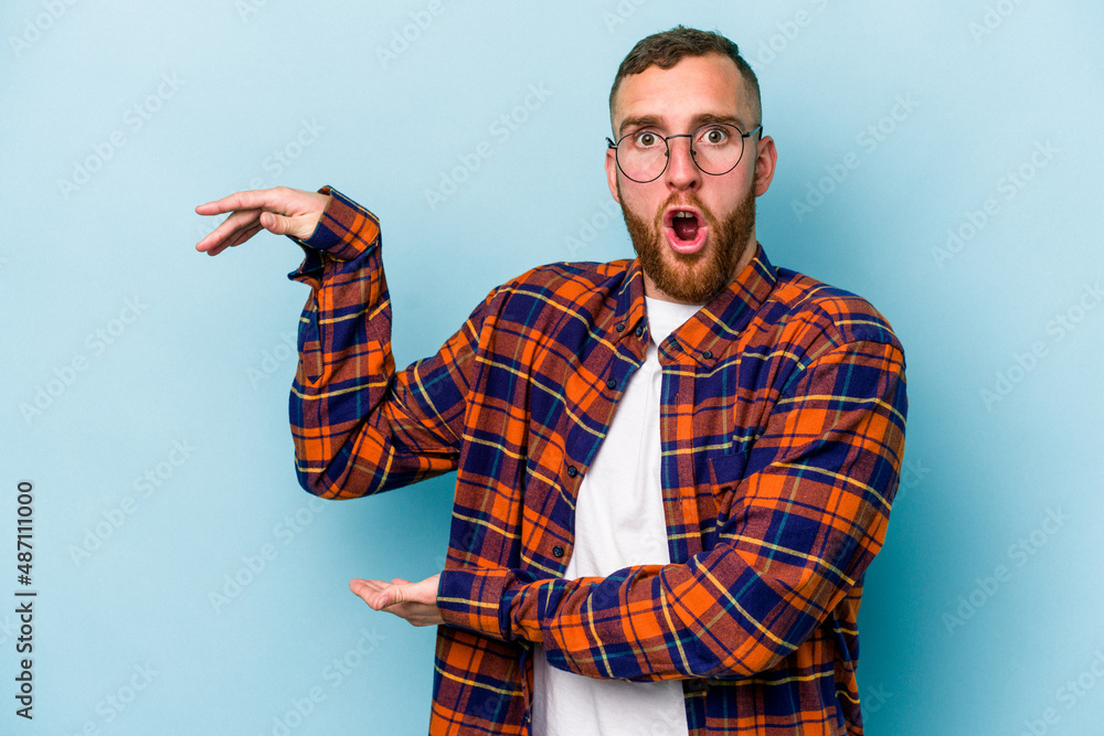 Young caucasian man isolated on blue background shocked and amazed holding a copy space between hands. Young caucasian man isolated on blue background shocked and amazed holding a copy space between hands.