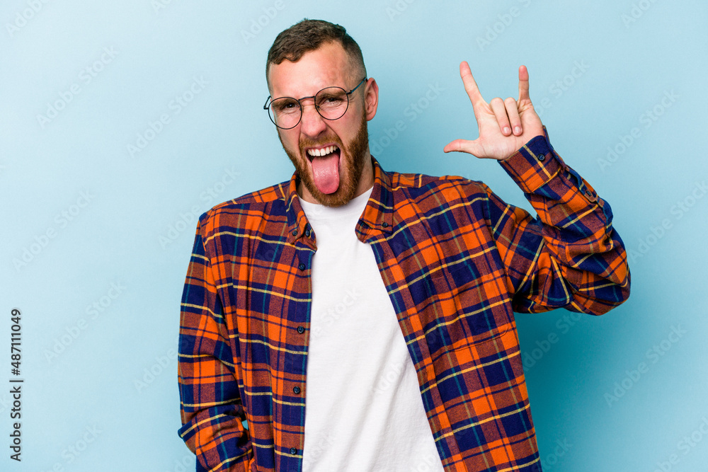 Naklejka premium Young caucasian man isolated on blue background showing a horns gesture as a revolution concept.