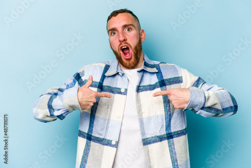 Young caucasian man isolated on blue background surprised pointing with finger, smiling broadly.