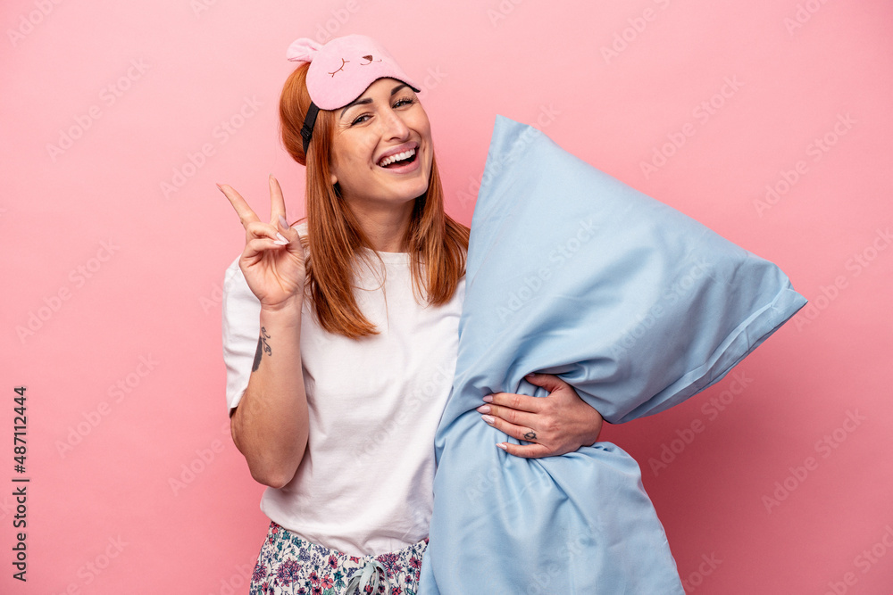 Young caucasian woman wearing a pajama holding pillow isolated on pink background joyful and carefree showing a peace symbol with fingers. Young caucasian woman wearing a pajama holding pillow isolated on pink background joyful and carefree showing a peace symbol with fingers.