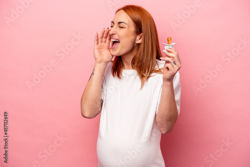 Young caucasian pregnant woman holding pacifier isolated on pink background shouting and holding palm near opened mouth.