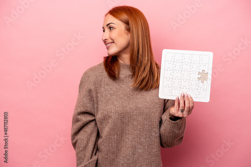 Young caucasian woman holding puzzle isolated on pink background looks aside smiling, cheerful and pleasant.