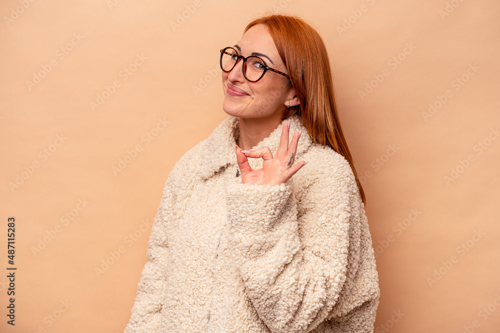 Young caucasian woman isolated on beige background winks an eye and holds an okay gesture with hand.