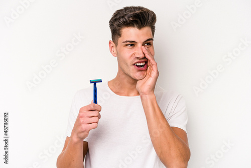 Young caucasian man shaving his beard isolated on white background is saying a secret hot braking news and looking aside