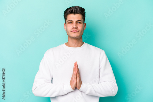 Young caucasian man isolated on blue background praying, showing devotion, religious person looking for divine inspiration. © Asier