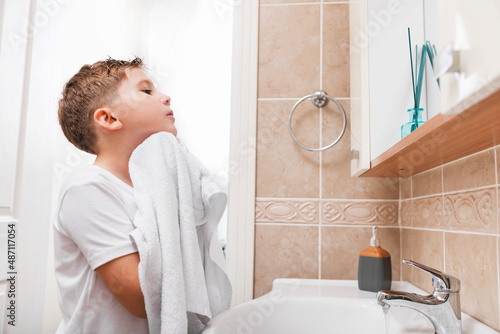 Health care, morning routine, wellness. Cute preteen boy wiping his face with a towel in the bathroom.