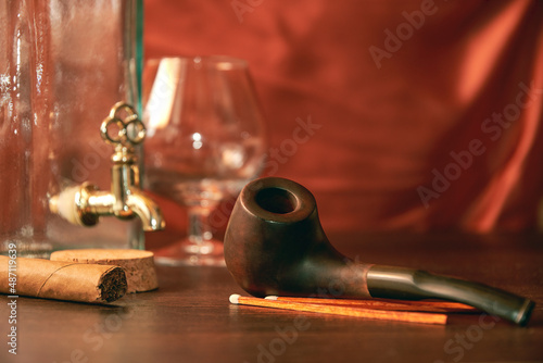 Smoking pipe with empty brandy glass and bottle and tobacco on the wooden table. Vintage man's relaxation stuff