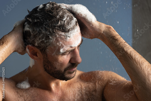 Handsome young man taking shower, washing his hair and body, standing under falling water drops in modern bathroom at home. Selfcare and wellness