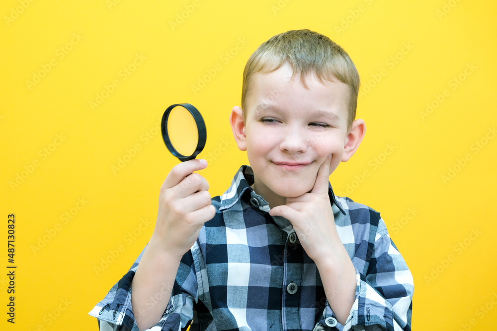 Positive curious schoolboy in casual clothes looks at the camera through a magnifying glass, on a yellow background. Positive curious schoolboy in casual clothes looks at the camera through a magnifying glass, on a yellow background.