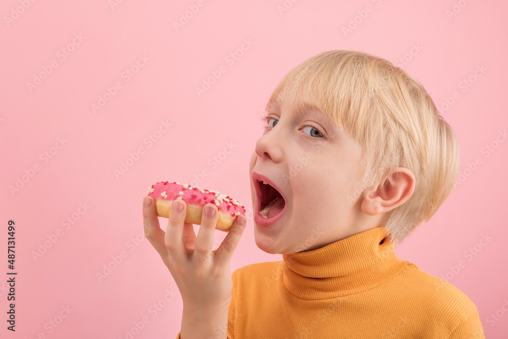 Fair-haired boy wants to bite donut with pink icing. Child has sweet tooth looks at the camera. Portrait of boy with bun on pink background. Fair-haired boy wants to bite donut with pink icing. Child has sweet tooth looks at the camera. Portrait of boy with bun on pink background.