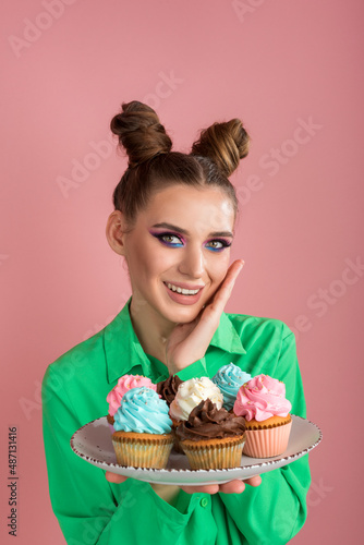 Playful girl with two bun hairstyle holds plate of cakes. Beautiful woman with bright makeup cupcakes on pink background. Vertical frame