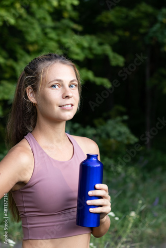 Vertical photo of a smiling sportive girl with a sports bottle of water outdoors on a blurred background of trees and forest. Woman in sportswear outdoors with a bottle of water.