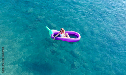 Young pretty woman in a white one piece swimsuit relaxing on a inflatable mattress in shape of big eggplant in turquoise sea water. Woman on air mtatress in transparent water. © Klemenso