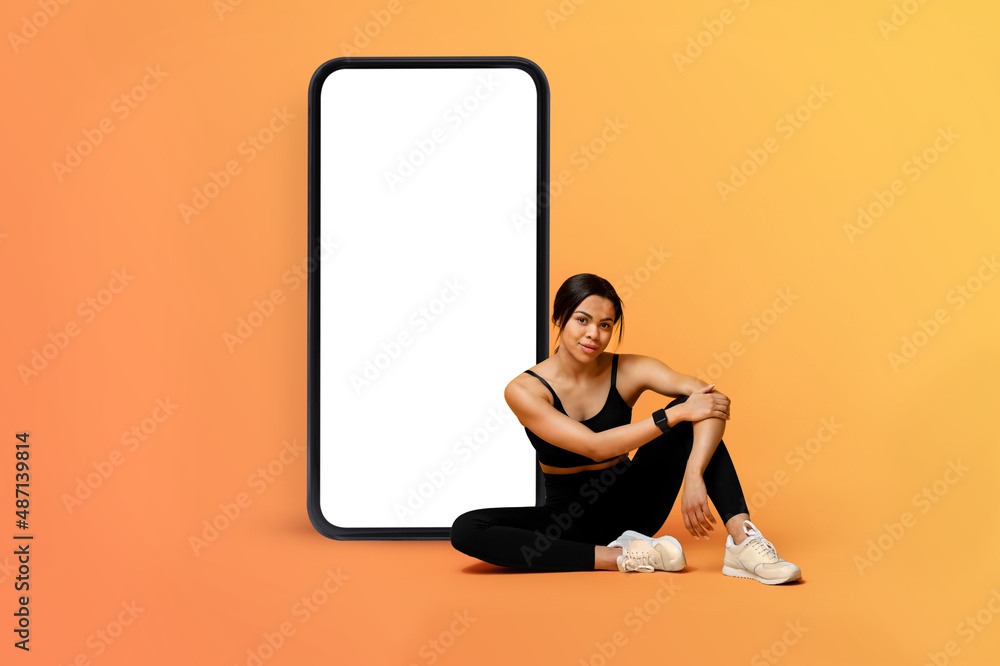 Young fitness lady sitting near huge smartphone with blank screen, demonstrating mobile app for online workout - obrazy, fototapety, plakaty 