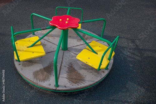 Wooden children carousel on anti-slip surface divided into six sectors with three yellow double seats and flower on central rotation axis. On handrails and floor traces of scuffed and peeling paint.