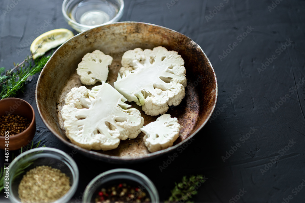 Cauliflower steak cooking. Raw cut cauliflower lie in a frying pan. Olive oil, herbs, various spices nearby. Dark background. Place for text.