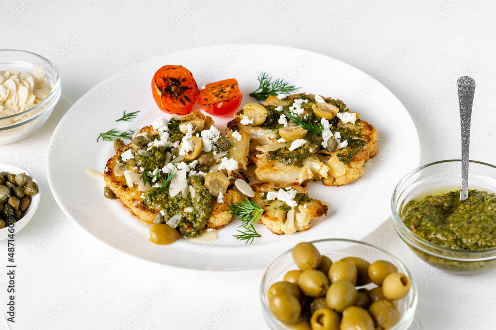 Cauliflower steak with spices, chimichurri sauce, almond flakes, olives, fried cherry tomatoes and capers on a white plate. Vegetarian food. White background. Cauliflower steak with spices, chimichurri sauce, almond flakes, olives, fried cherry tomatoes and capers on a white plate. Vegetarian food. White background.