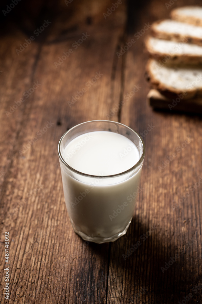 A full glass of milk. Bread in the background is blurred. Dark wooden background. Morning breakfast. Healthy food. Vertical frame. A full glass of milk. Bread in the background is blurred. Dark wooden background. Morning breakfast. Healthy food. Vertical frame.