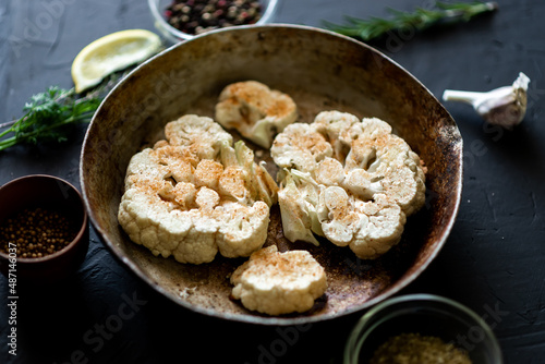 Cauliflower steak cooking. Raw cauliflower sprinkled with spices lies in a frying pan. Olive oil, herbs, various spices nearby. Dark background.