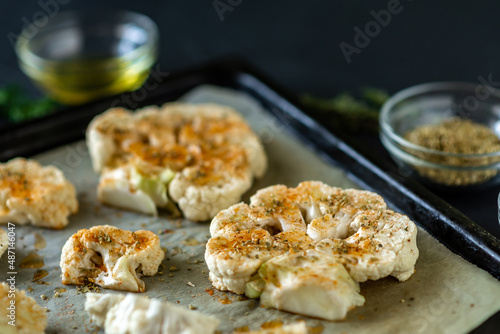 Cauliflower steak cooking. Raw cauliflower sprinkled with spices lies on a baking sheet. Olive oil, herbs, various spices nearby. Dark background.