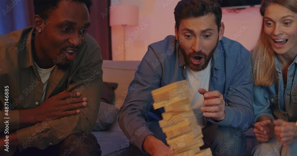 Cheerful Group of Friends of Men and Girls Playing Game Jenga at Home. Happy Friends Have Fun playing with Wooden Blocks in living room. While their friend accidentally Falls Down the Jenga Tower.