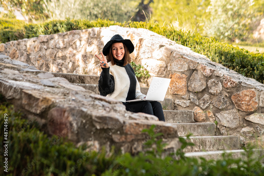 Una linda y alegre mujer caucásica con un sombrero 
sentada en las escaleras de un parque trabajando con su ordenador