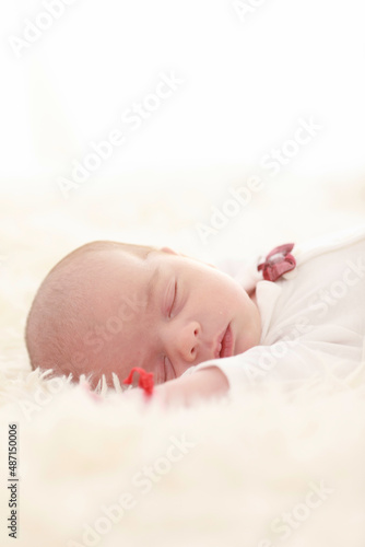 

7 day old baby sleeps happily in bed on a fluffy white blanket dressed in a white shirt and red bow tie in a white room