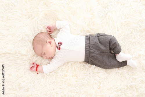 

7 day old baby sleeps happily in bed on a fluffy white blanket dressed in a white shirt and red bow tie in a white room