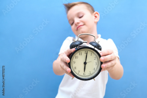 a boy on a blue background with glasses holds an alarm clock in his hands. it is difficult for a child to wake up in the morning